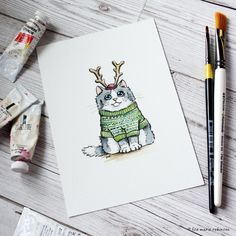 a card with a cat wearing a sweater and reindeer antlers on it, next to paintbrushes