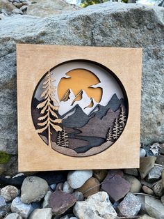 a wooden plaque with mountains and trees in the background on some rocks, along with pebbles