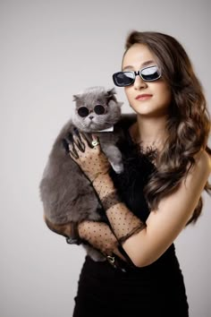 a woman in black dress holding a grey cat wearing sunglasses and lace gloves on her arm