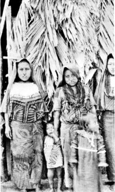 an old black and white photo of three people standing in front of a hut with thatched roof