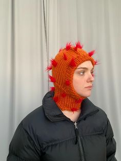 a man wearing a knitted hat with red spikes on it's head, standing in front of a curtain