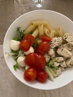 a white bowl filled with pasta, tomatoes and lettuce next to a glass of wine