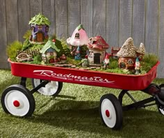 a red wagon filled with lots of small houses on top of green grass next to a wooden fence