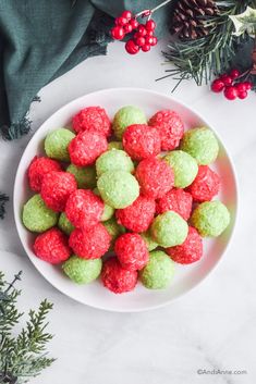 a white bowl filled with green and red snowball cookies next to evergreen branches, pine cones and berries