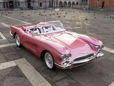 an old pink car parked in front of a building with pigeons on the ground around it