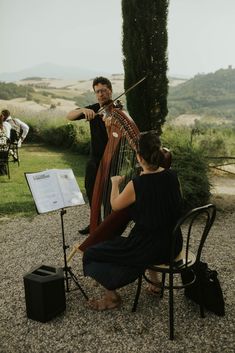 two people are playing music on the grass near a tree and an outdoor area with chairs