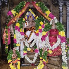 the statue is surrounded by flowers and garlands on it's head, with two men sitting in front of him