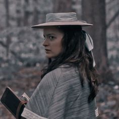 a woman wearing a hat and holding a book in her hands while walking through the woods