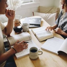 two people sitting at a table with open books and coffee mugs on the table