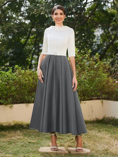 a woman standing in the grass wearing a long gray skirt and white top with sandals