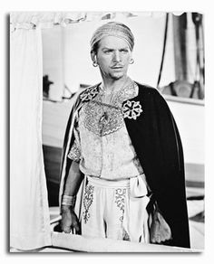 a black and white photo of a man dressed in traditional clothing standing next to a boat