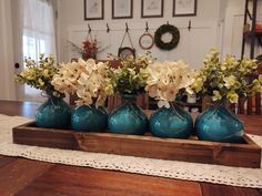 four blue vases with flowers in them sitting on a wooden tray at the end of a dining room table