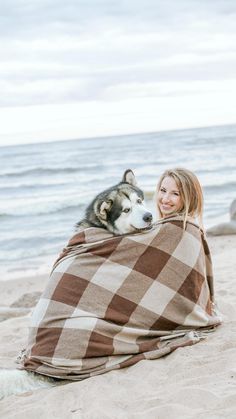 a woman wrapped up in a blanket on the beach with her husky dog laying next to her