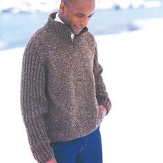 a man standing in the snow wearing a brown sweater and blue pants with his hand on his hip