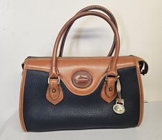 This vintage Dooney & Bourke satchel top handle bag is a stylish addition to any wardrobe. The navy blue/beige exterior color and pebbled finish give it a classic 80s/90s designer look. The bag features a zip closure, double handles, and a bag charm accessory.  With a size of 12 inches in width, 8 inches in height, and 6 inches in depth, it is a medium-sized bag suitable for various occasions such as travel, weddings, parties, casual outings, and workwear. The bag is made of high-quality leather Vintage Double Handle Satchel For On-the-go, Vintage Bags With Gold-tone Hardware And Double Flap, Vintage Satchel Tote With Gold-tone Hardware, Navy Leather Shoulder Bag With Gold-tone Hardware, Blue Vintage Bag With Gold-tone Hardware, Medium Sized Bags, Dooney Bourke, Leather Purse, High Quality Leather