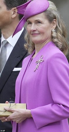 a woman in a purple dress and hat standing next to a man wearing a suit