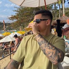 a man with tattoos on his arm drinking from a cup while standing under an umbrella