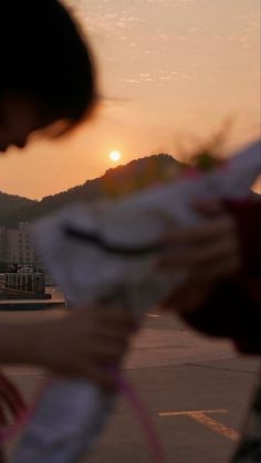 a woman is holding something in her hand and looking down at it with the sun setting behind her