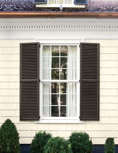 an open window with black shutters on a white brick building in front of green bushes