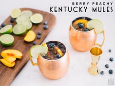 two copper mugs filled with blueberries, lime and peaches on top of a cutting board