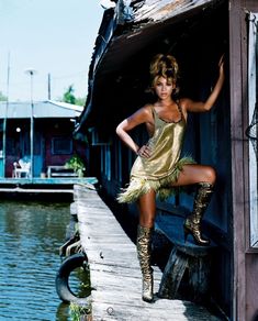 a woman in a gold dress standing on a dock with her legs crossed and wearing boots