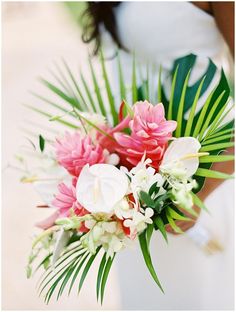 a bride holding a bouquet of pink and white flowers in her hand with palm leaves