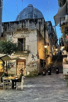 two people sitting at an outdoor table with umbrellas in the middle of a cobblestone street