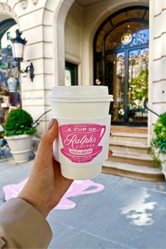 a person holding up a cup of coffee in front of a building with pink flowers