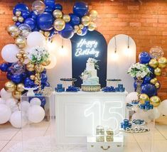 a table topped with cake and balloons in front of a brick wall filled with blue, gold and white decorations