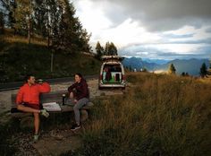 two people sitting on a bench next to a van in the mountains with trees and grass