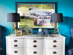 a white dresser with two lamps and pictures on it in front of a blue wall