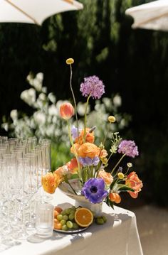 an arrangement of flowers, fruit and wine glasses on a white table cloth with umbrellas in the background