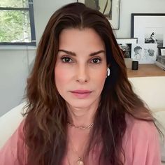 a woman with long brown hair is looking at the camera and she's wearing a pink shirt