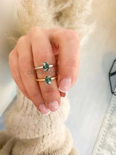 a woman's hand holding an engagement ring with green stones on it, while wearing a white sweater