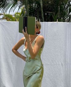 a woman in a green dress holding a book up to her face while standing next to a white wall