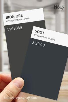 The image compares two deep, dark paint colors. On the left, "Iron Ore" by Sherwin Williams (SW 7069) is displayed in a rich, dark gray tone with bold black text above it. On the right, "Soot" by Benjamin Moore (2129-20) is shown in a very dark blue-black tone with similar bold black text. The swatches are angled and held by a hand against a blurred indoor background, highlighting the differences between these two dramatic, dark shades. Elegant Backdrop, Trim Colors, Best White Paint, Types Of Colours, Iron Ore, Color Scale, Types Of Lighting, Trim Color, Coordinating Colors