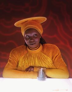 a woman sitting at a table with a cup in front of her and an orange background behind her