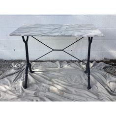 a white marble table with black iron legs on a tarp covered ground in front of a brick wall