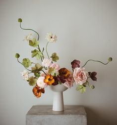 an arrangement of flowers in a white vase on a concrete block against a gray wall