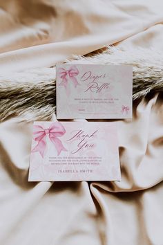 two cards with pink bows on them sitting on a white bed sheet in front of a fur