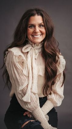 a woman with long brown hair wearing a white shirt and black pants, smiling at the camera