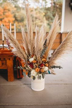 an arrangement of dried flowers and grasses in a white vase on a table with candles