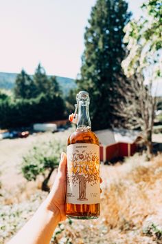 a person holding up a bottle of alcohol in front of a field with trees and buildings