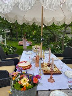 an outdoor table set up for a party with cake and flowers on the table under an umbrella