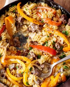 a skillet filled with pasta and vegetables on top of a wooden table next to a napkin