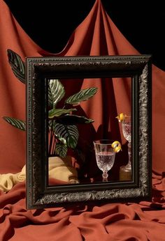 a framed photograph with two wine glasses and a potted plant in the reflection on a red cloth