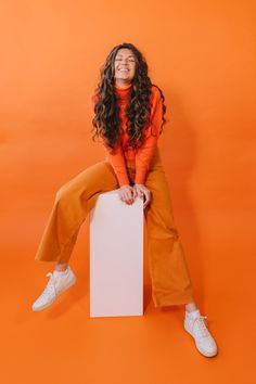 a woman sitting on top of a white box in front of an orange wall smiling