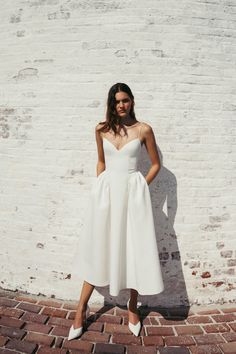 a woman standing in front of a brick wall wearing a white dress with bow at the waist