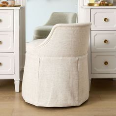 an upholstered chair sits in front of a white dresser with gold knobs