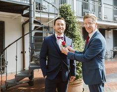 two men standing next to each other in front of a building and one is tying his bow tie